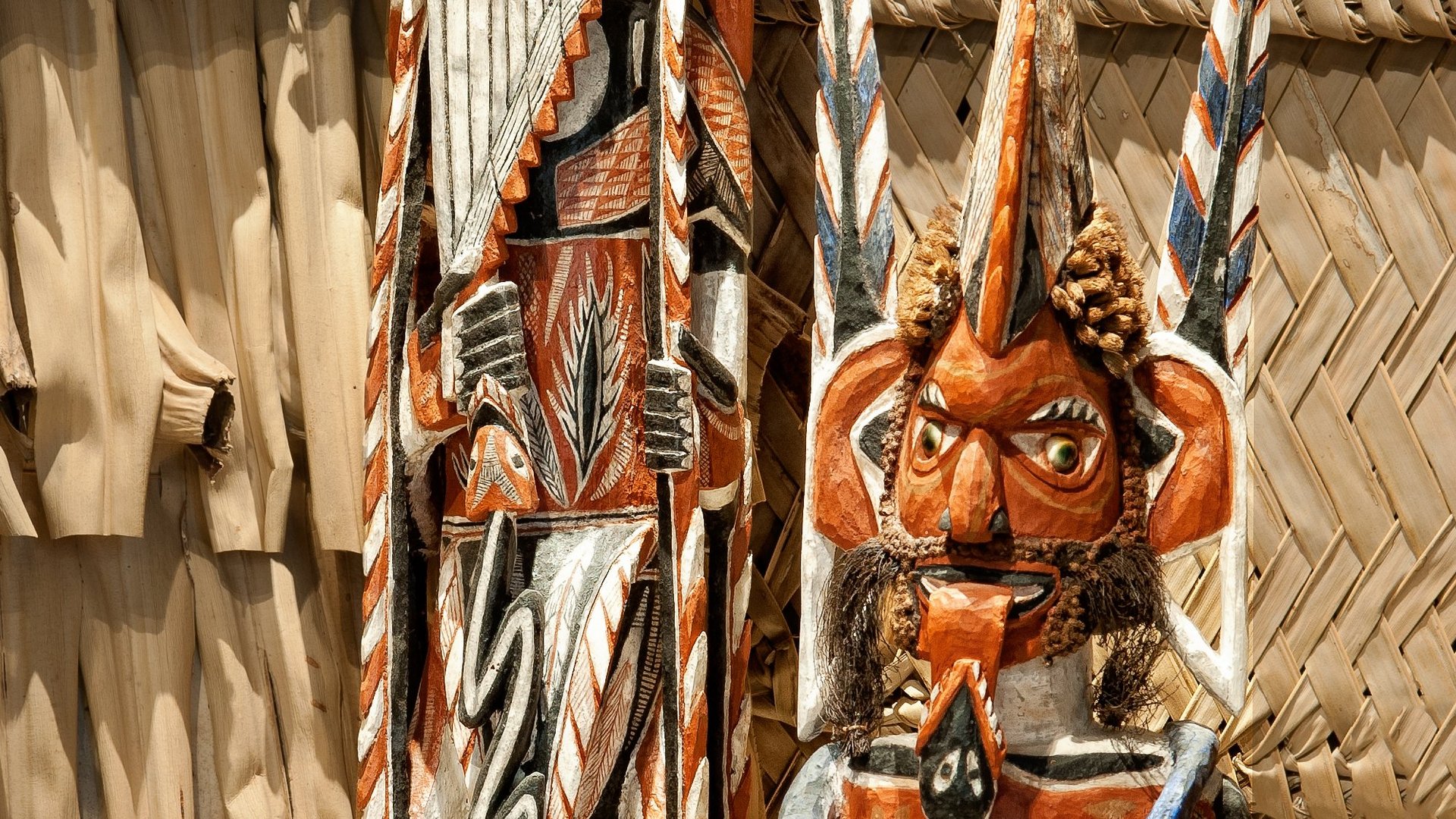 Photo: MUT | Valentin Marquardt Three painted carvings in column form, depicting stilised persons with one carrying a spear