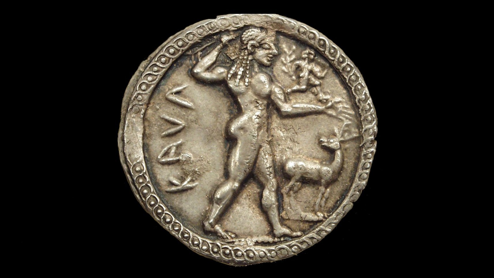 Obverse of a silver coin depicting a young Apollo walking to the right; his arm ist stretched out and has a small, male figure running on it; to the god's feet a stag turns its head to him; to the left of Apollon is the Greek inscription of "KAUL" (short for Kaulonia)