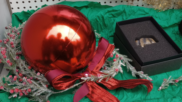 Foto: MUT | Nadja Mozdzen Auf grünem Tuch liegt zur Linken eine rote Christbaumkugel mit roter Schleife auf einem Kranz aus falschem Immergrün, zur Rechten liegt ein wenig zurückgesetzt ein schwarzes Schächtelchen mit der Replik der Mammutfigur aus Elfenbein