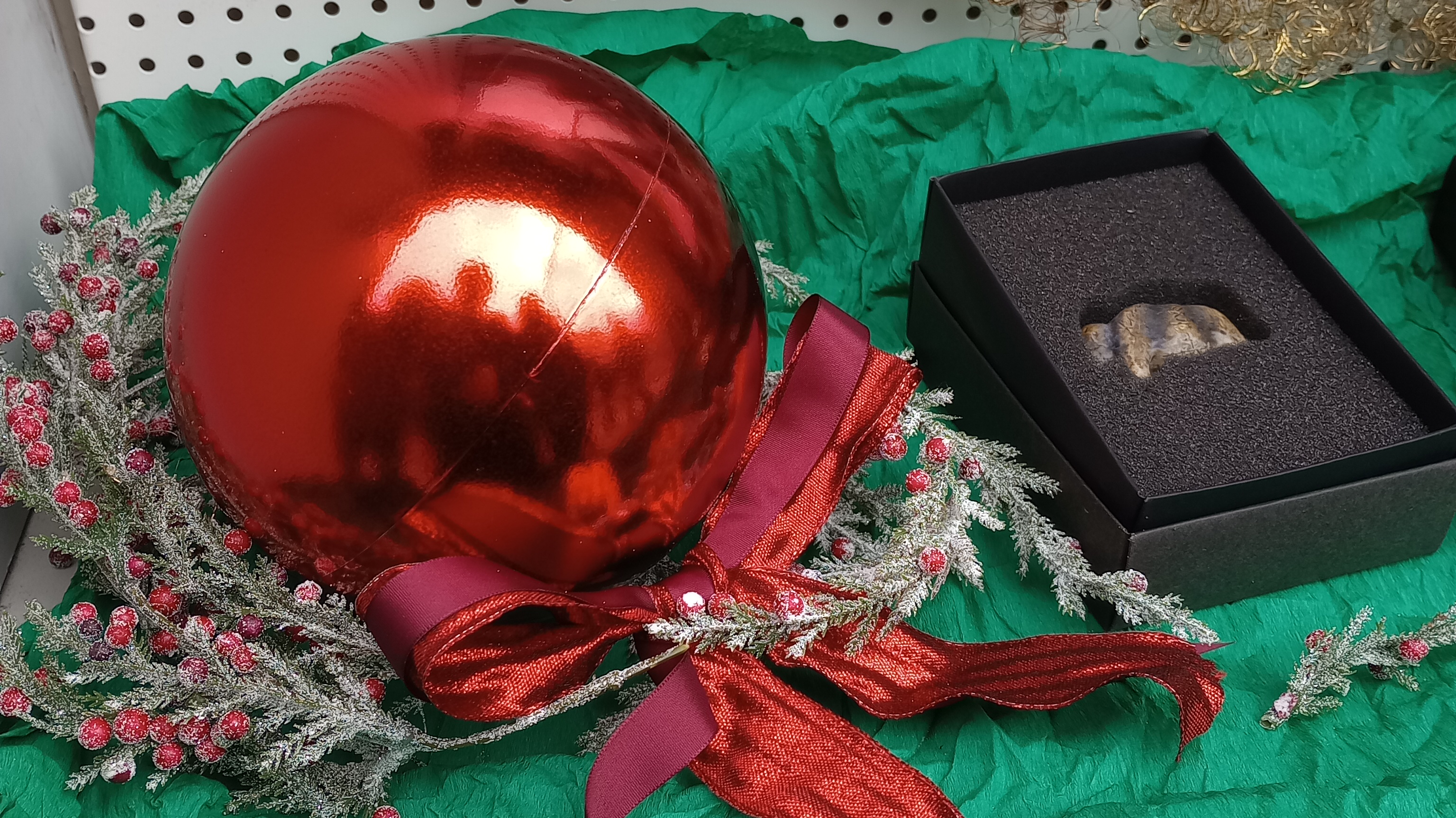 Auf grünem Tuch liegt zur Linken eine rote Christbaumkugel mit roter Schleife auf einem Kranz aus falschem Immergrün, zur Rechten liegt ein wenig zurückgesetzt ein schwarzes Schächtelchen mit der Replik der Mammutfigur aus Elfenbein