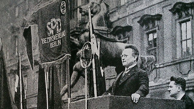 Foto in schwarz-weiß: Paul Löbe steht auf einem Rednerpodest vor dem Berliner Schloss im Oktober 1930; neben ihm ein Banner der SPD.
