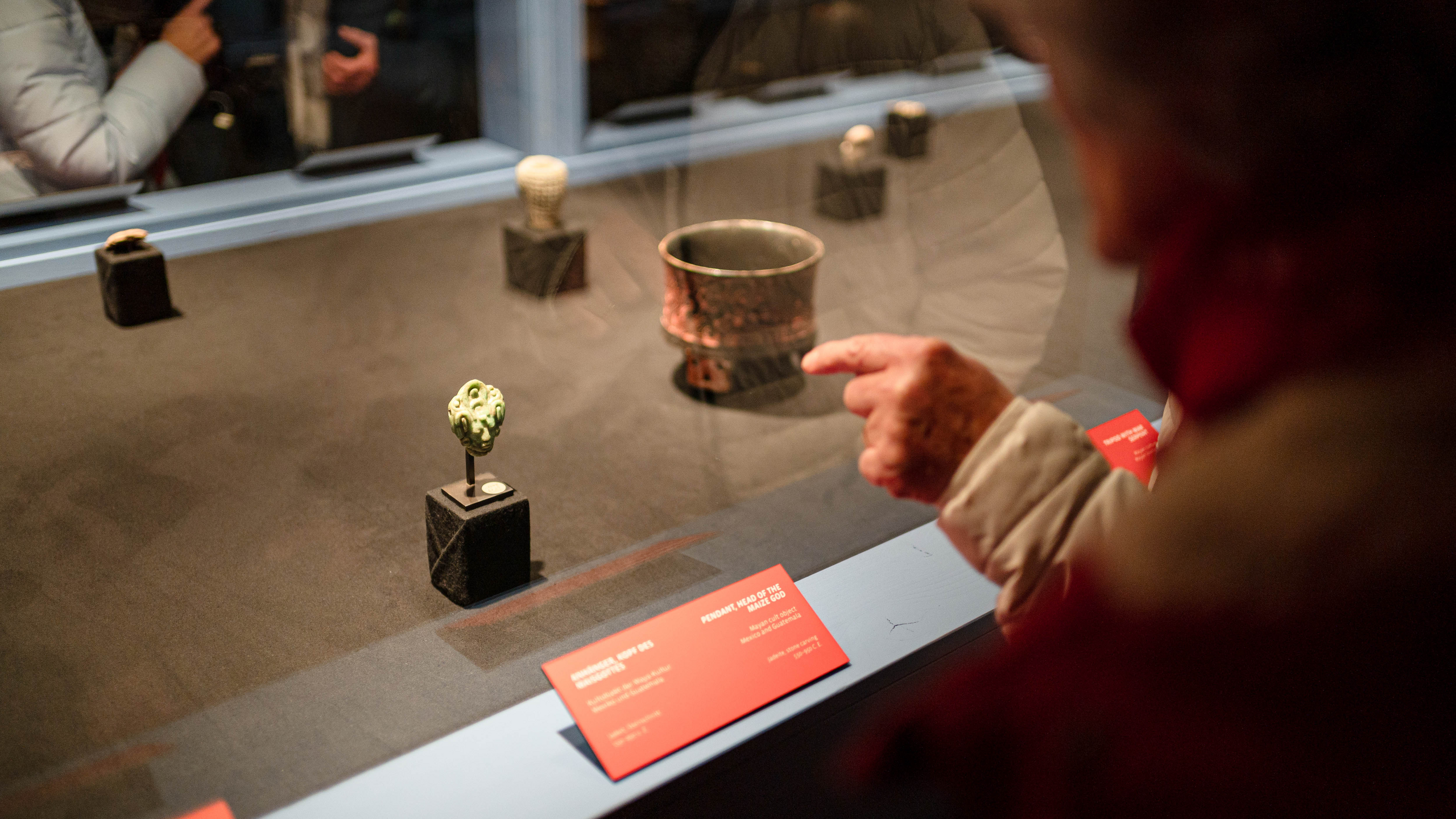 Blick in den maya-blauen Vitrinenkatafalk, eine nicht zu erkennende Person deutet durch die Scheibe hindurch auf einen Jadekopf, dessen Objektbeschriftung am Rande der Vitrine außen angebracht ist