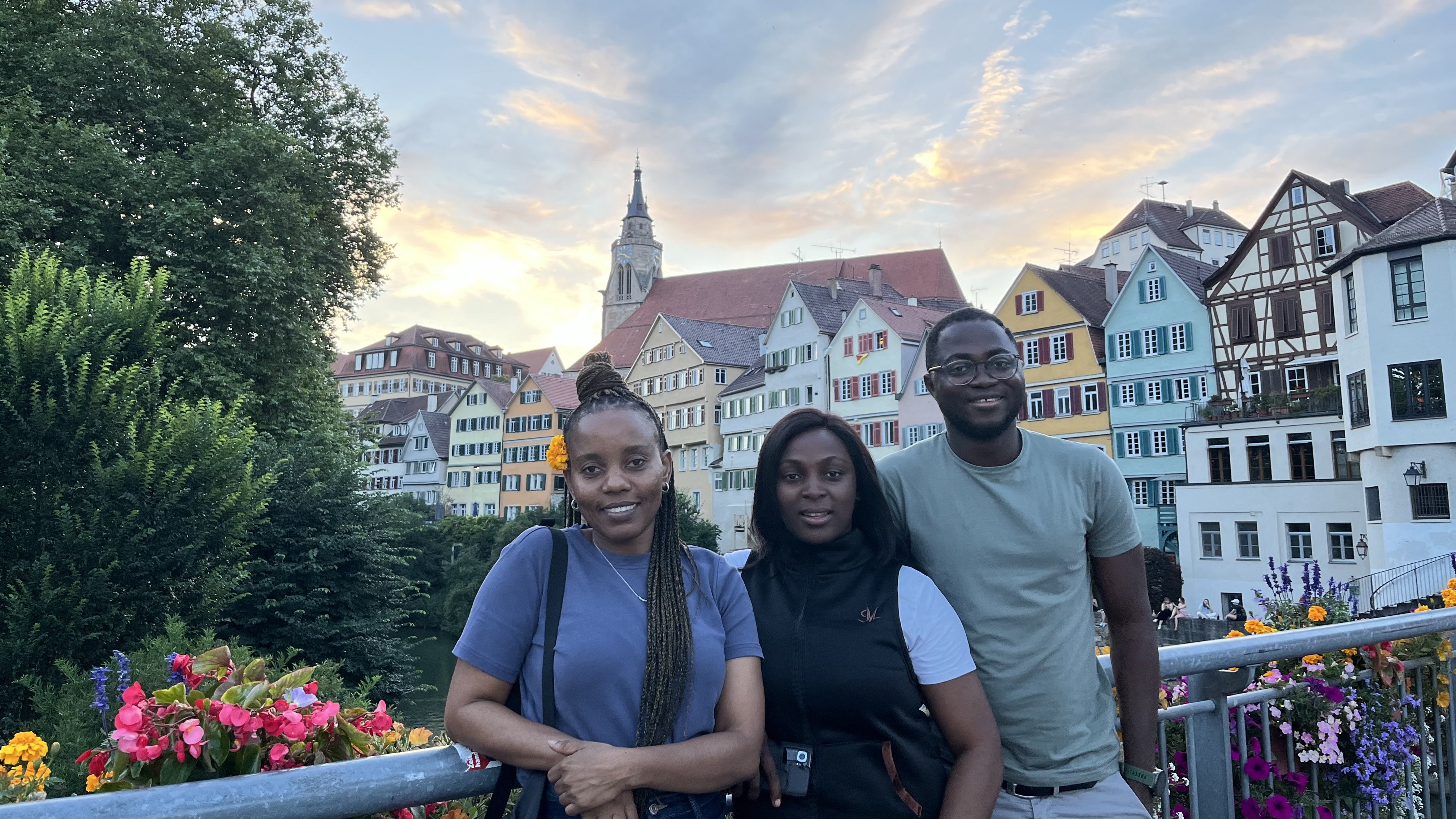 Drei Personen auf der Neckarbrücke in Tübingen. Im Hintergrund sind Bäume, bunte Blumen und Häuser zu sehen.