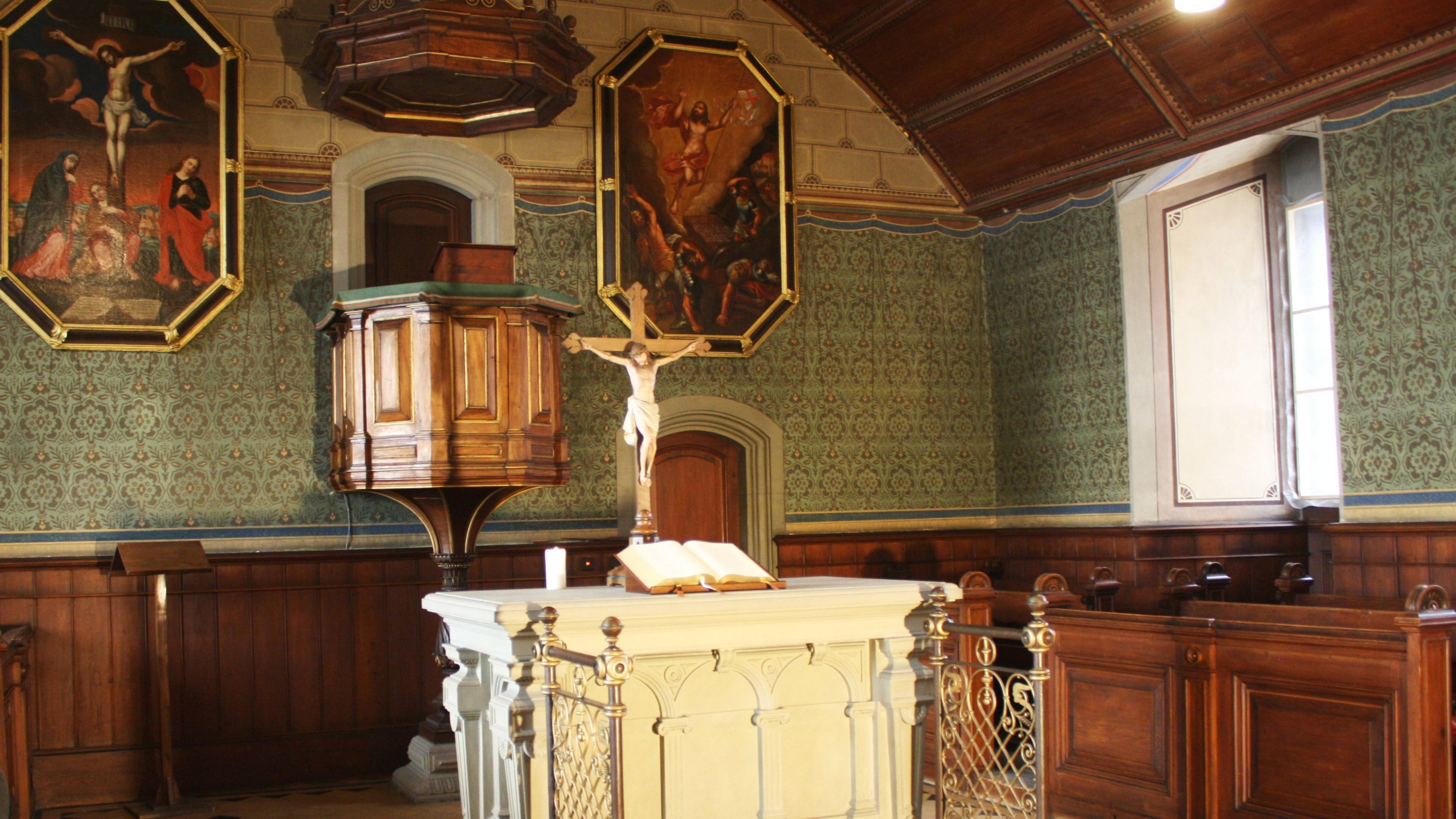 View of the altar area of the castle church, with the white altar and the opened bible and Christus figure upon it; in the background is the pulpit, flanked by tall painting depicting the crucified (left) and ascending (right) Christus respectively