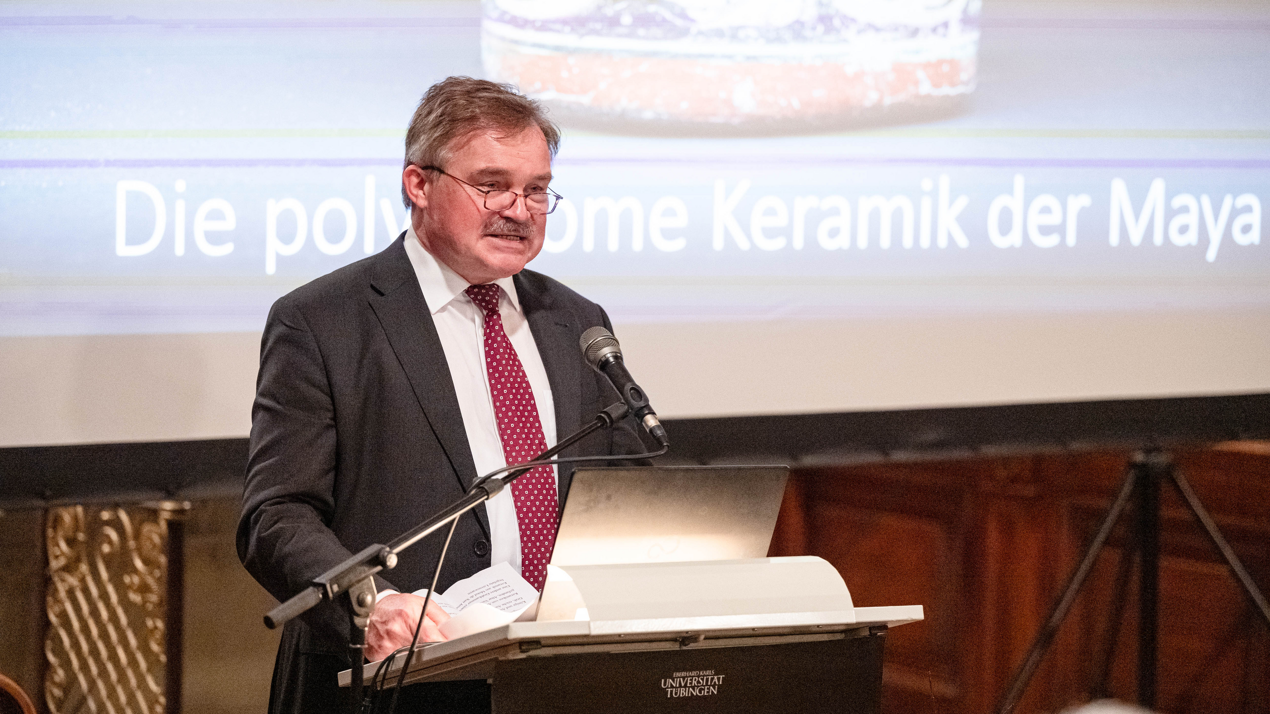 Professor Dr. Dr. h. c. Nikolai Grube at the lectern in front of the altar of Hohentübingen Castle church in front of the screen on which the picture of a Mayan ceramic mug and the title "Die polychrome Keramik der Maya" (Mayan polychrome ceramics) is projected