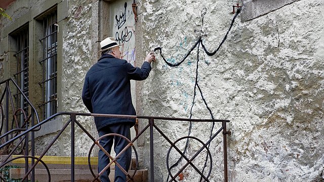 Foto: Charlotte Hermann und Uli Metz Treppenabsatz an einem Steingebäude, auf dessen Fassade ein älterer Herr in weißem Hut und dunkler Jacke eine simplistische Figur sprüht