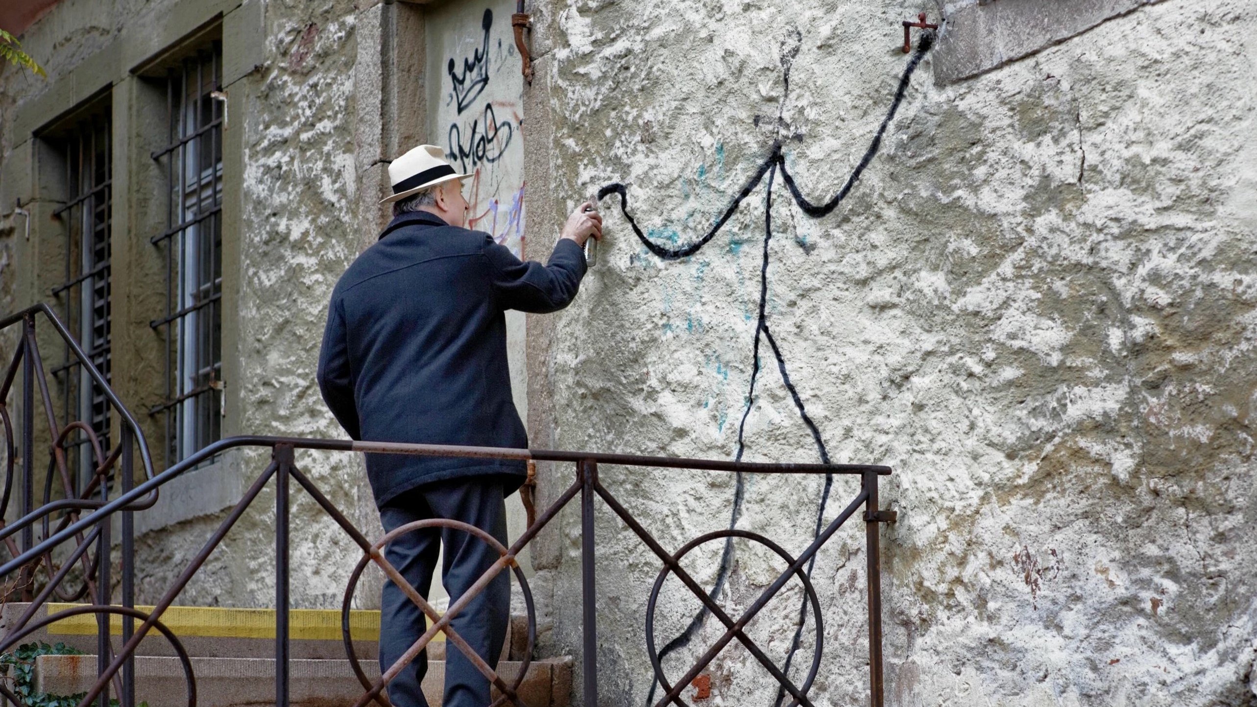 Treppenabsatz an einem Steingebäude, auf dessen Fassade ein älterer Herr in weißem Hut und dunkler Jacke eine simplistische Figur sprüht