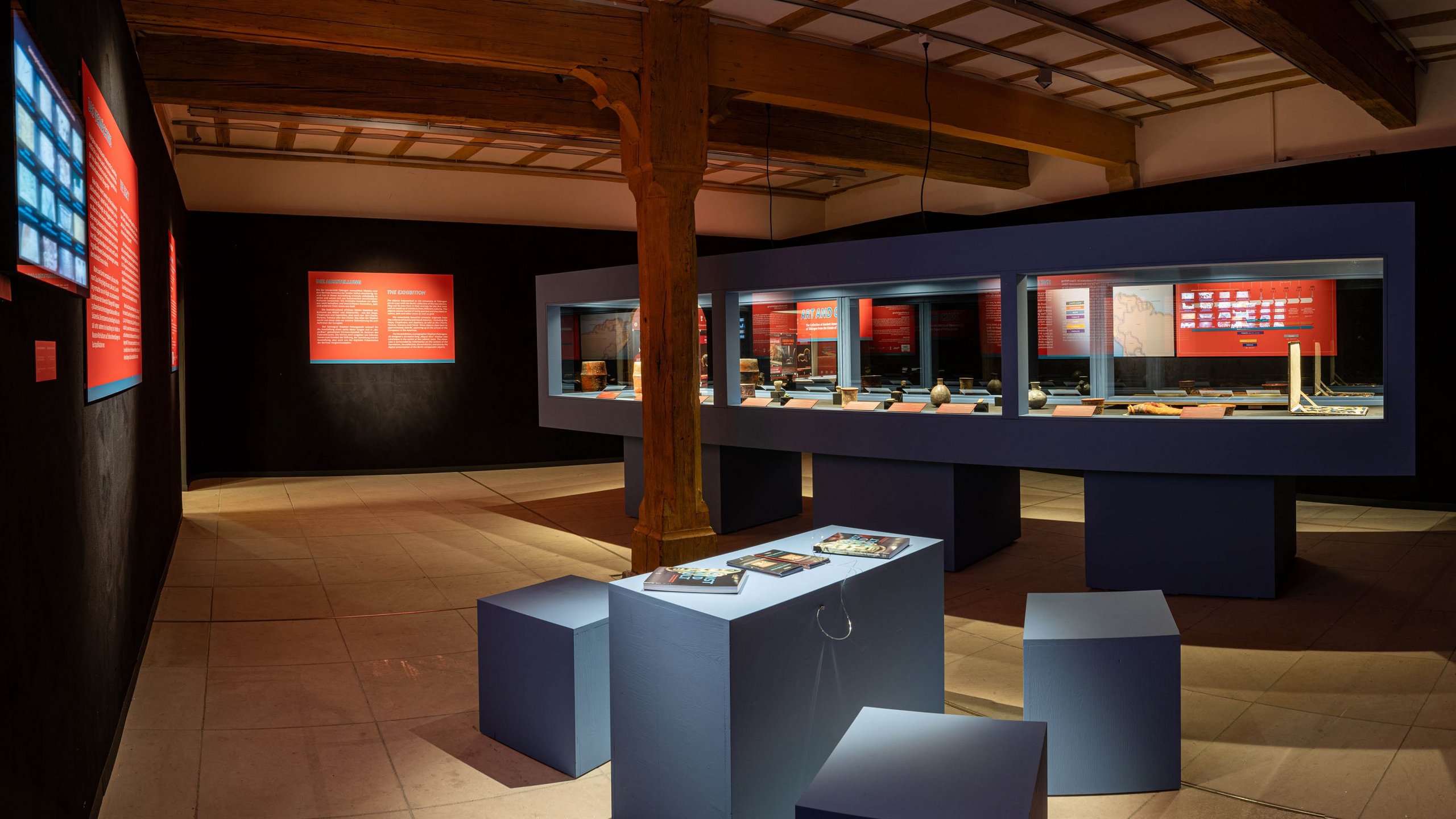 Blick in den menschenleeren Ausstellungsraum; vor dem schräg stehenden, beleuchteten Vitrinenkatalfalk steht eine Sitzgruppe aus Maya-blauen Kuben, auf denen der Katalog "Kunst und Kult" angekettet ausliegt