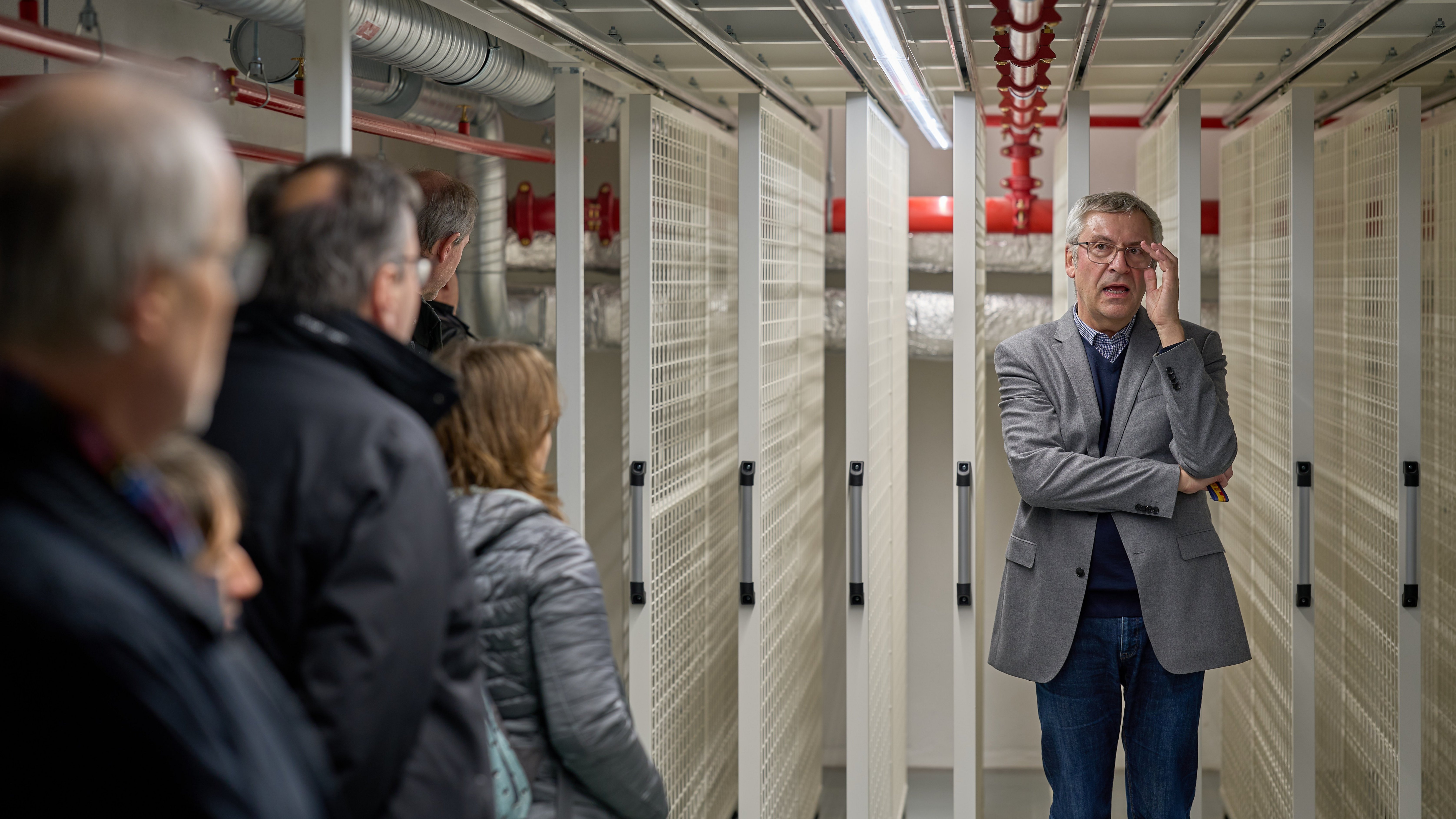 Ernst Seidl steht in erklärendem Gestus vor den noch leeren Aufhängegittern für die Gemäldesammlung; vorne links sind unscharf die ihn Betrachtenden