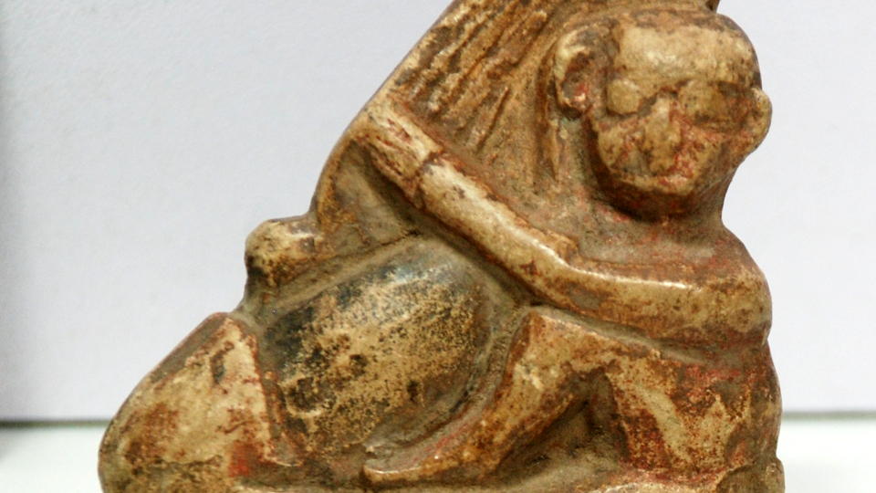 Kleinformatige Steinfigur eines kahlen Mannes (rechts), der einen übergroßen Phallus (links) umarmt
