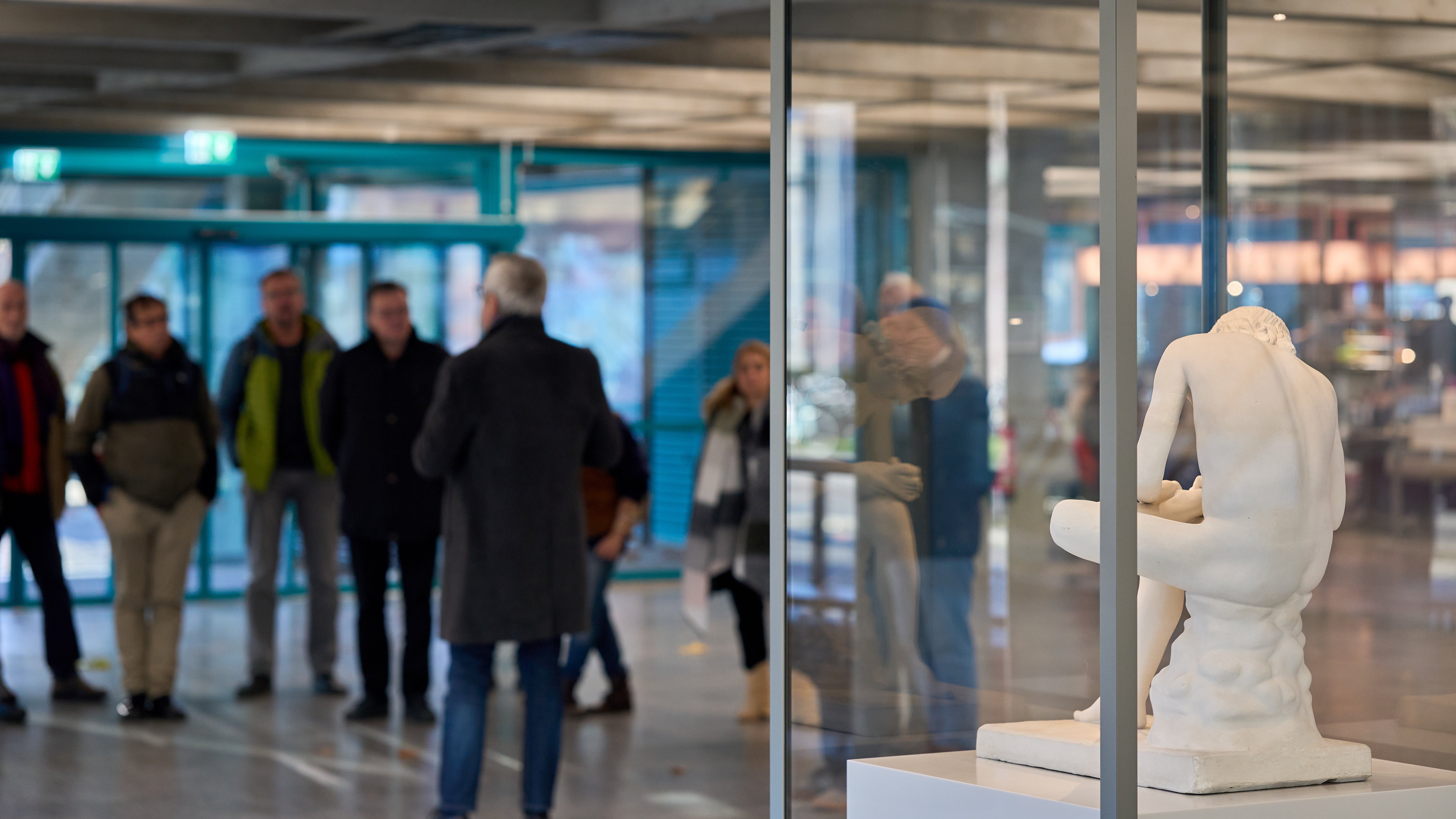 Eine Gruppe aus Männer und Frauen steht zuhörend dem Direktor im Foyer der Mensa gegenüber; vorne rechts als einziges im Fokus ist die Rückansicht der Schauvitrine mit einer sich einen Dorn aus dem Fuß ziehenden, antiken Statue