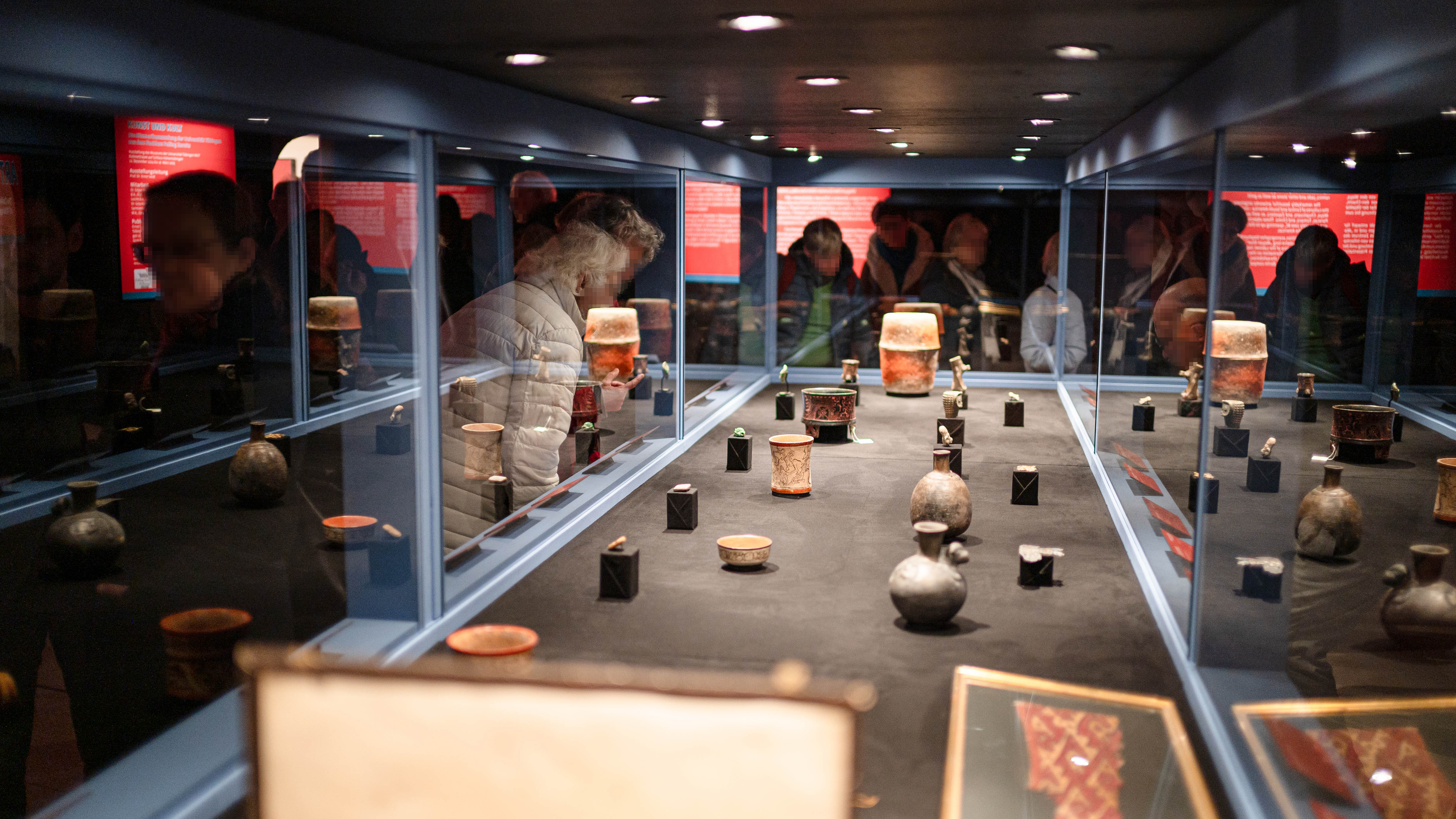 Blick längst durch eine lange, beleuchtete Vitrine, die mit Exponaten aus Keramik, Jade und anderem Stein bestückt ist; durch die spiegelnden Glasflächen sind die umstehenden Menschen zu sehen, deren Gesichter verschwommen oder verpixelt sind