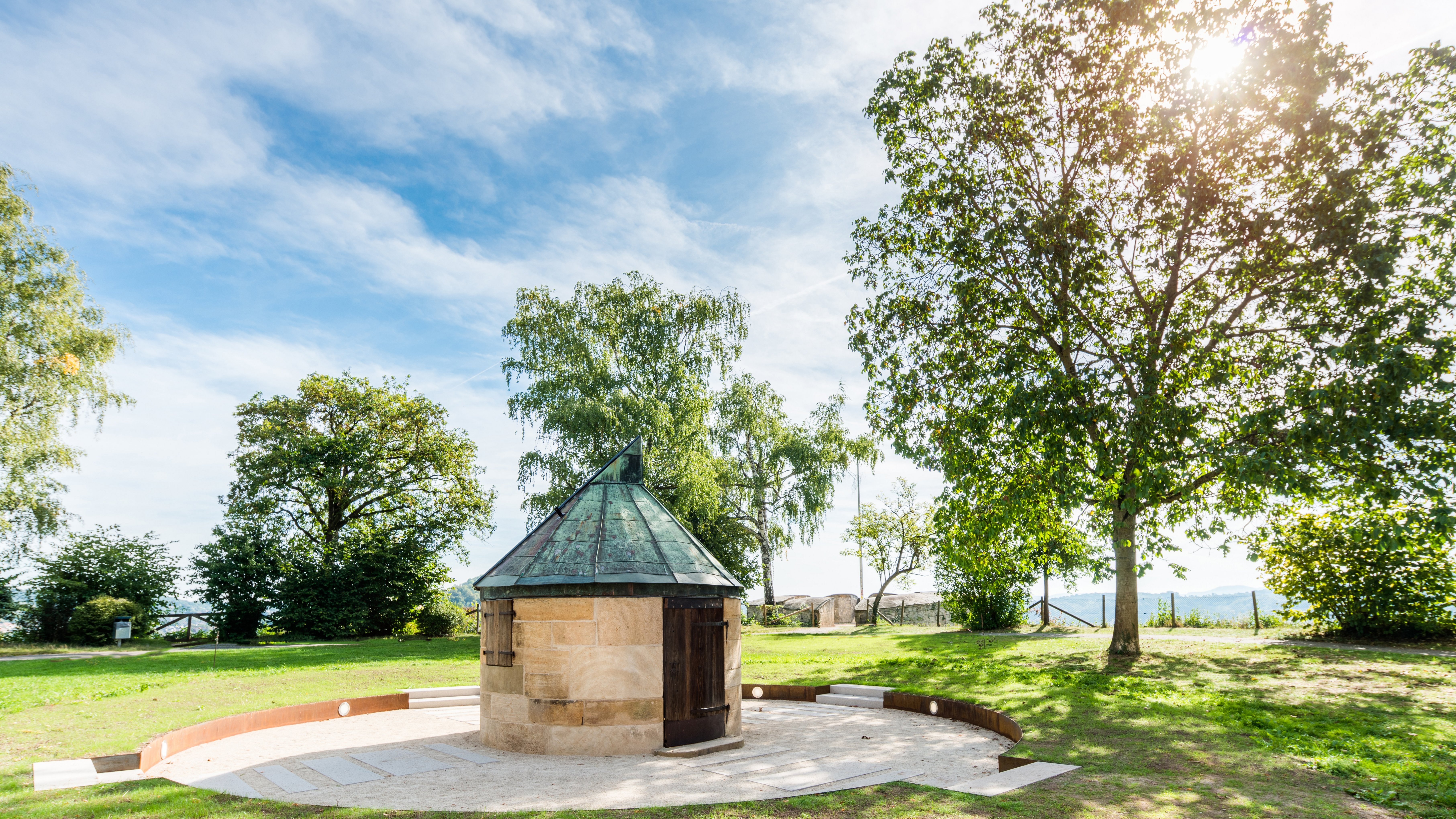 Small circular building with copper roof standing on a paved round, with green grass and trees around it