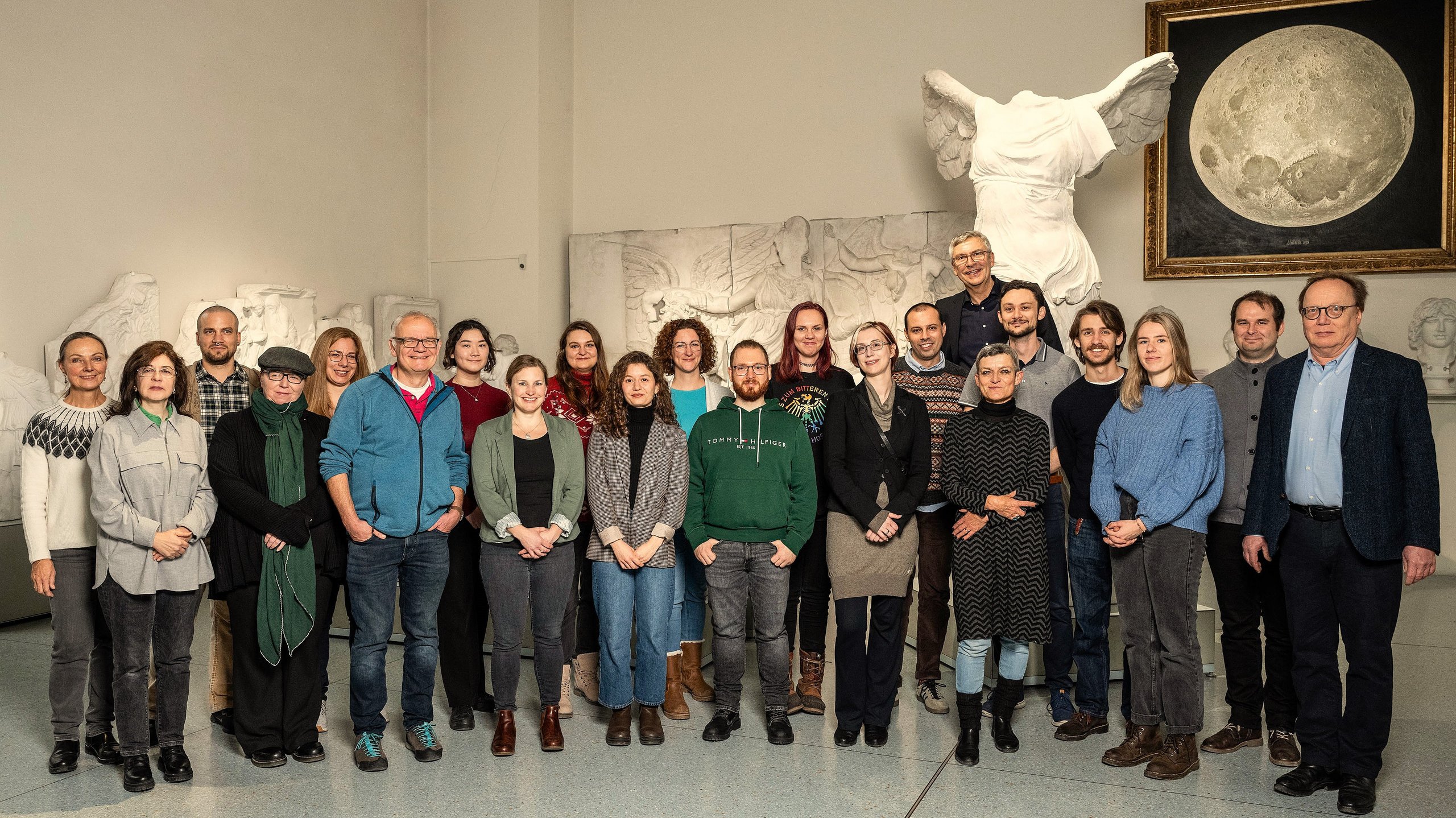 Teamfoto der MUT-Mitarbeitenden im Halbkreis im Rittersaal auf Schloss Hohentübingen; im Hintergrund der Gipsabguss der Nike von Samothrake und das realistische Mond-Gemälde