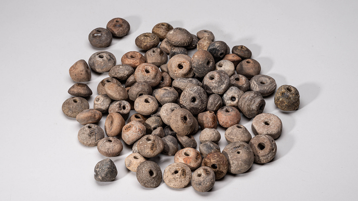 KLA-OR-14, Bronzezeitliche Spinnwirtel und Webgewichte Spinnwirtel und Webgewichte mit eingeritzten Linien, in der Bildmitte angehäuft, vor neutral-grauem Hintergrund.