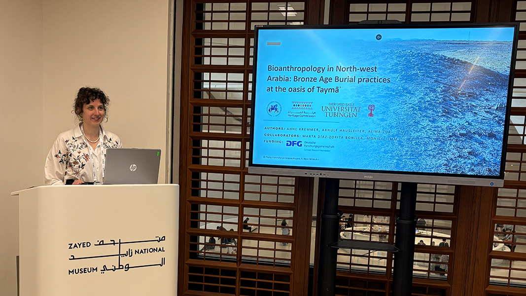58th Seminar for Arabian Studies, Abu Dhabi Dr. Anne Kremmer am weißen Rednerpult mit ihrer Präsentation mit dem Titel "Bioanthropology in North-west Arabia: Bronze Age Burial practices at the oasis of Taymā"" auf einem Bildschirm rechts