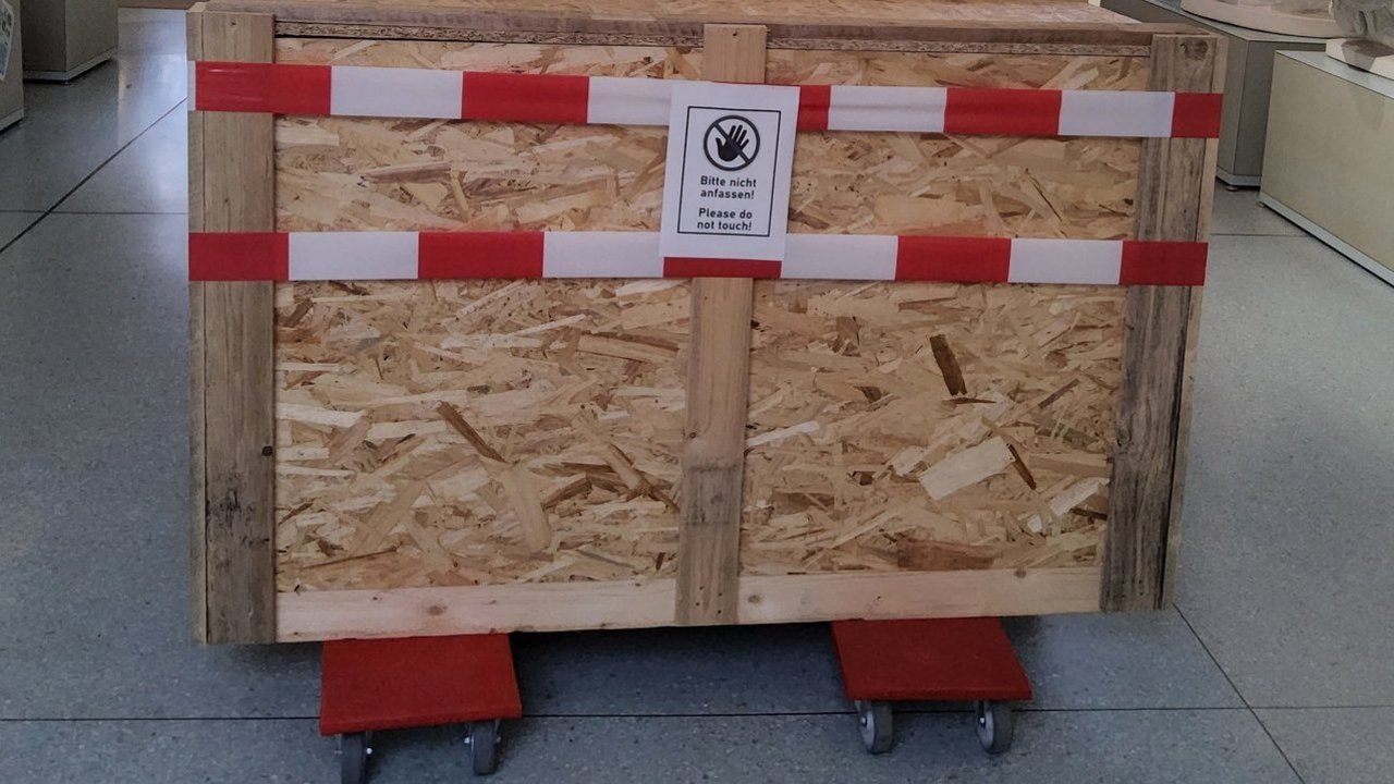 Eine Kiste auf Rollbrettern mit Absperrband umwickelt; ein aufgeklebtes Schild mit einem Handsymbol sagt "Bitte nicht anfassen! Please do not touch!"