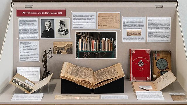 Frontansicht einer Vitrine mit aufgeschlagenen Büchern und Texten sowie Portraits von Max und Giuseppina Fleischmannauf der Rückwand