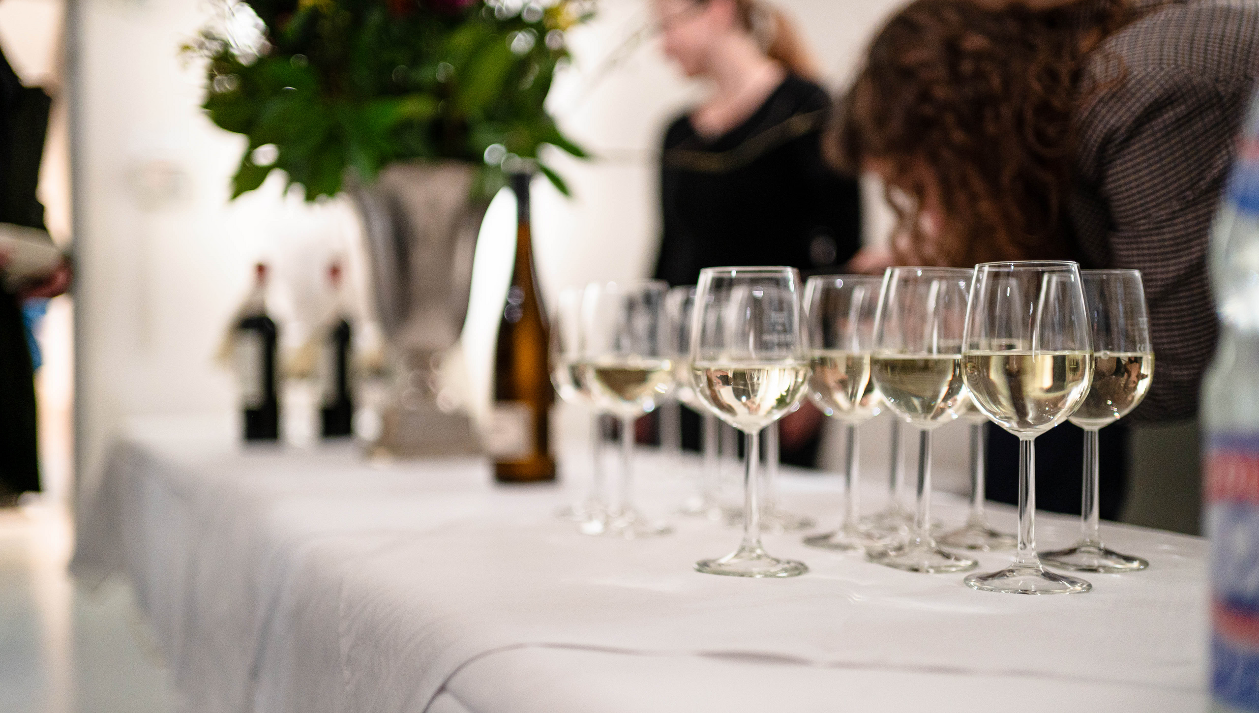 Flacher Blick über ein weiß gedecktes Buffet mit gefüllten Weißweingläsern und einem unscharfen Hintergrund, in dem ein Pokal mit Blumen steht