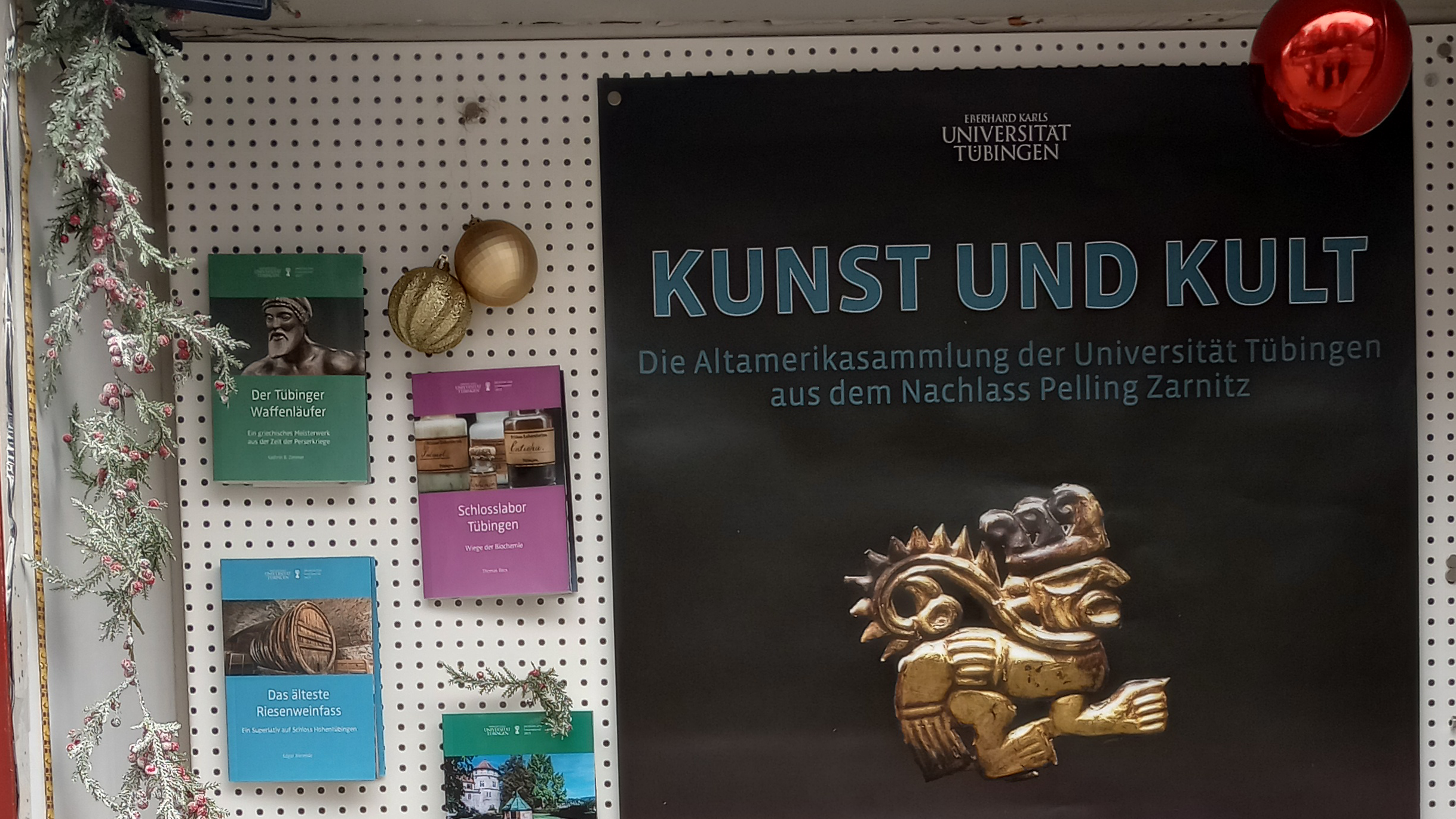 Rückwand der Vitrine, auf der links die kleinen Monographien und rechts das Poster zur Ausstellung "Kunst und Kult" mit einem goldenden, figürlichen Ornament angebracht sind; links hängt eine Kette aus falschem Immergrün