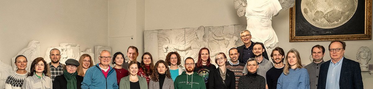 Teamfoto der MUT-Mitarbeitenden im Halbkreis im Rittersaal auf Schloss Hohentübingen; im Hintergrund der Gipsabguss der Nike von Samothrake und das realistische Mond-Gemälde