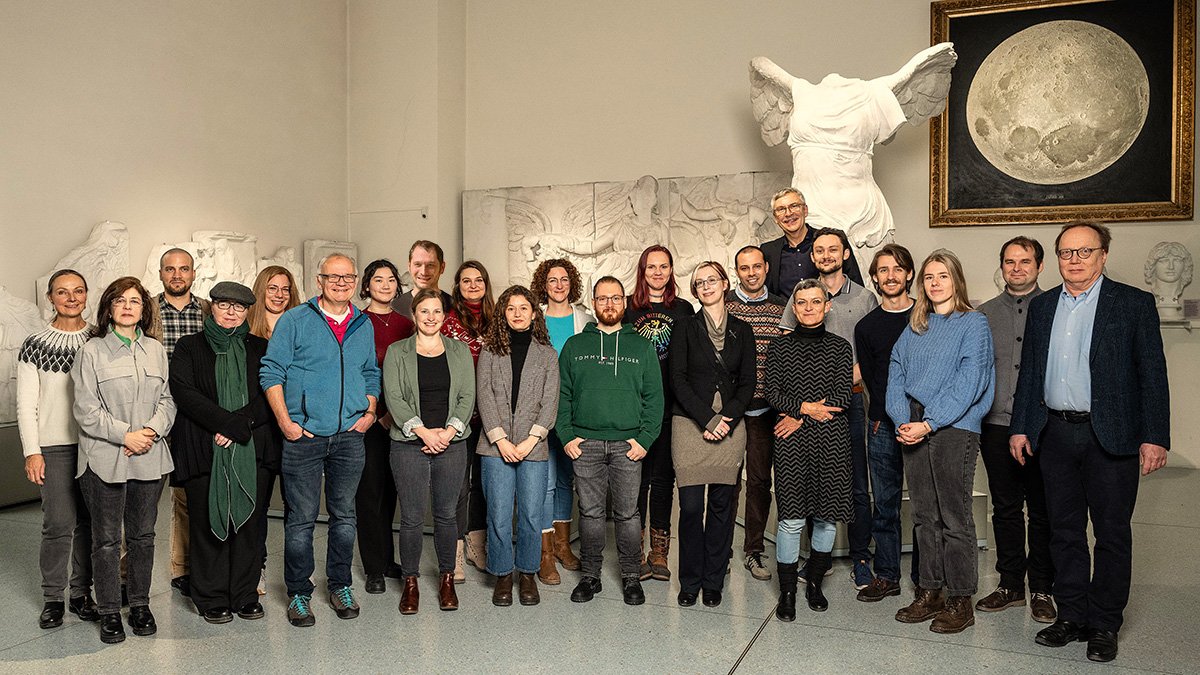 Teamfoto der MUT-Mitarbeitenden im Halbkreis im Rittersaal auf Schloss Hohentübingen; im Hintergrund der Gipsabguss der Nike von Samothrake und das realistische Mond-Gemälde