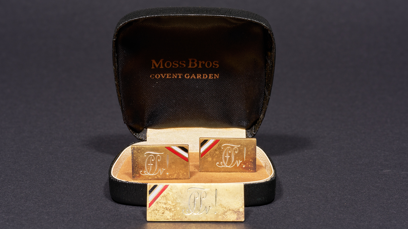 Geöffnete schwarze Schmuckschatulle mit einem paar goldener Manschettenknöpfe mit dem Sigil der Corps Suevia, vor der Schatulle eine rechteckige Krawattennadel mit demselben Motiv