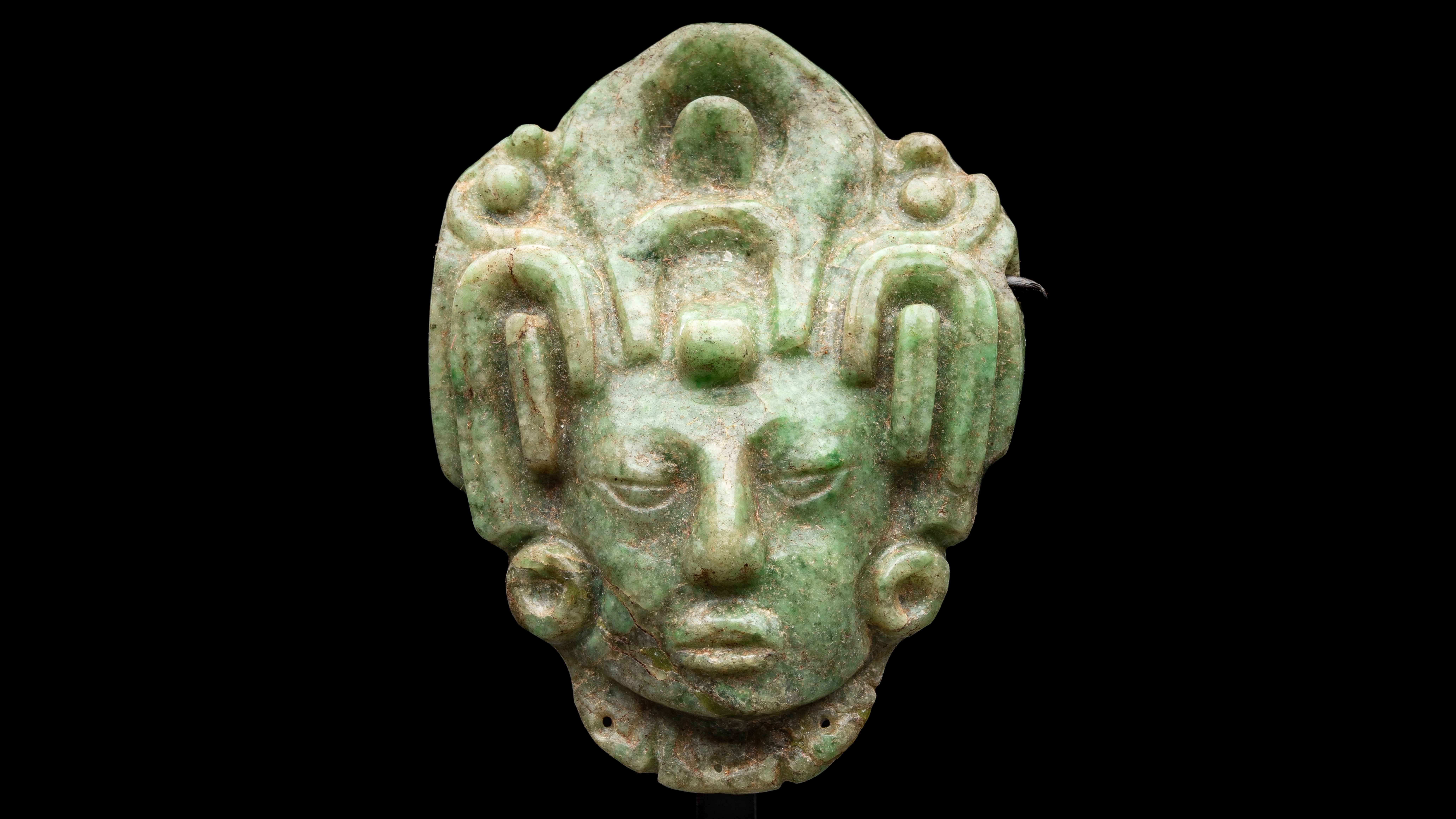 Green jade head of a Mayan maize god with headdress against a black background