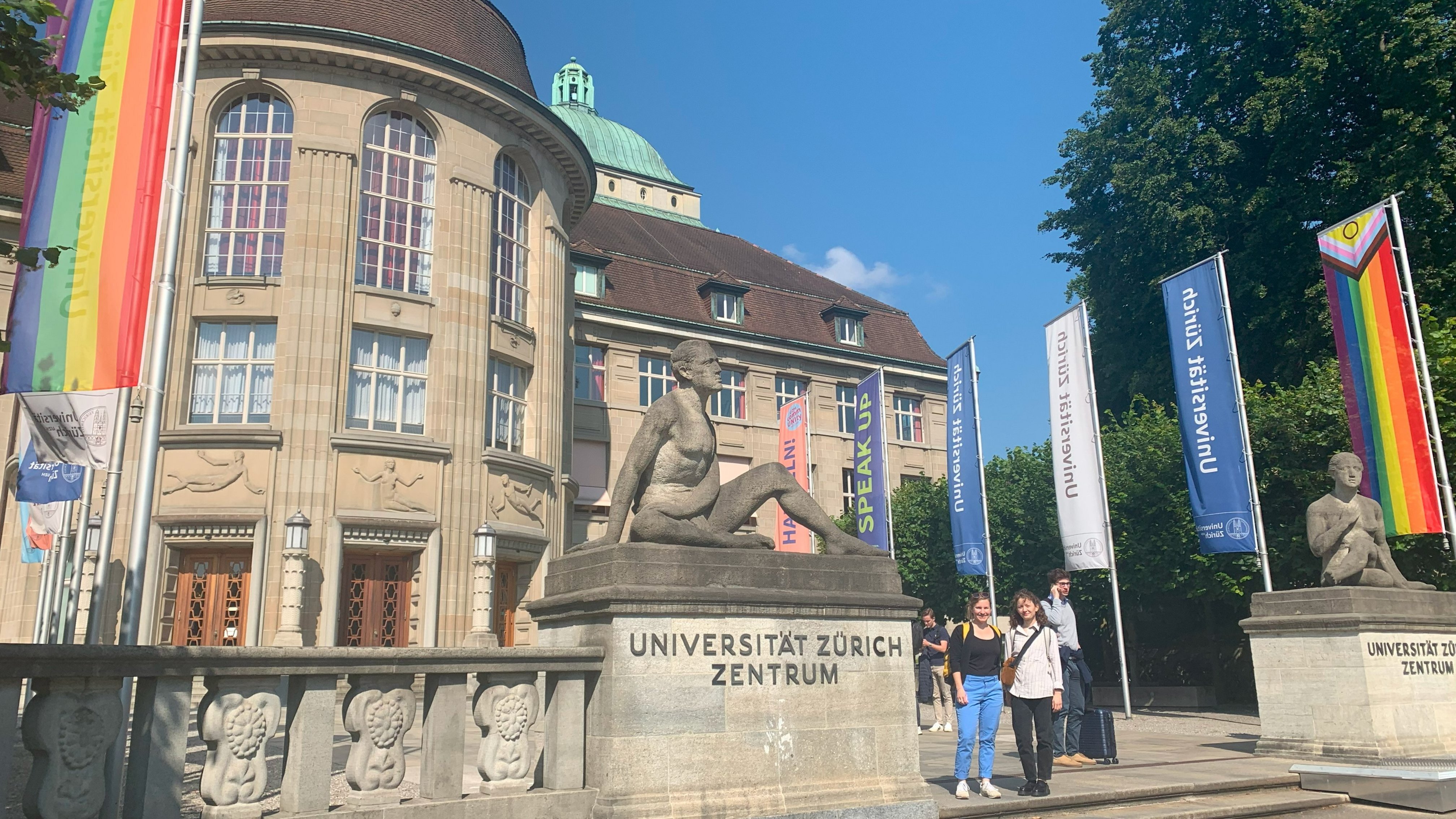 Zu sehen ist der Haupteingang der Universität Zürich mit einigen Flaggen und zwei Mitarbeiterinnen des MUT, Annika Vosseler und Diellëza Hyseni