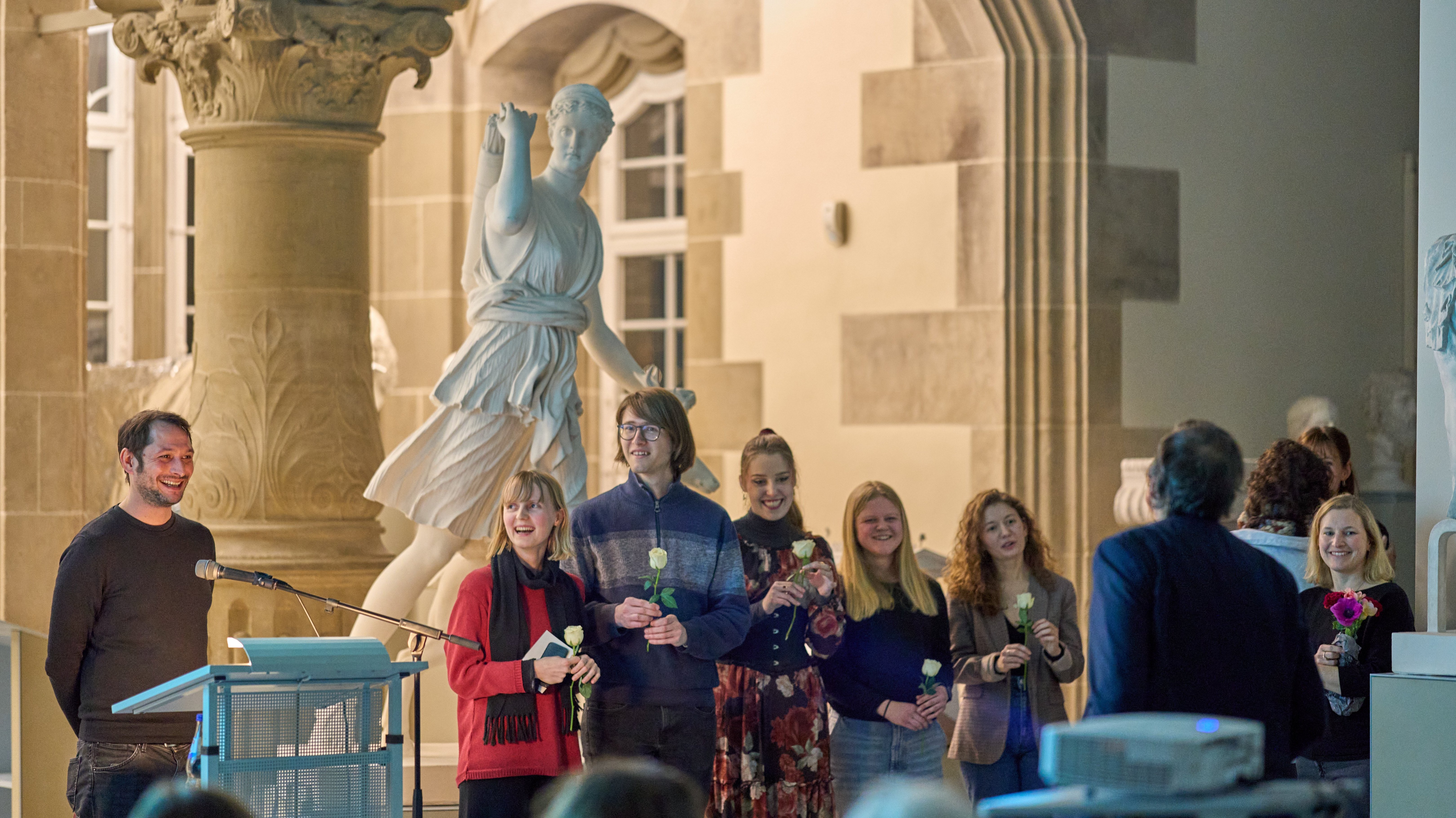 Am Rednerpult Dr Michael La Corte, daneben (v. l. n. r.) Solveig Schreiber, Timon Butz, Anastasia Rotenberg, Leyla Waberski, Diellëza Hyseni mit jeweils einer weißen Rose in der Hand; ganz rechts im Bild Dr des Annika Vosseler mit Blumenstrauß