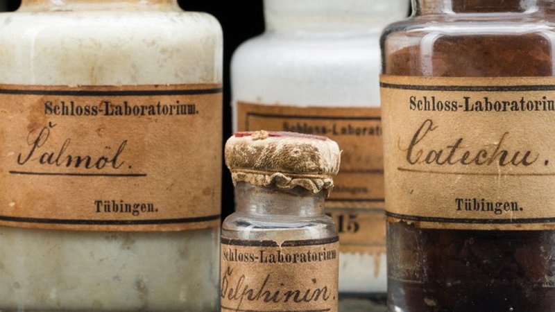 Ausschnitt eines Fotos mehrerer historischer Ingredienzfläschchen mit vergilbten Etiketten; Aufschrift jeweils "Schloss-Laboratorium" und "Tübingen" sowie auf einer Flasche mit weißer, fleckiger Masse "Palmöl", Fläschchen mit bräunlichen Pulver "Catechu" und kleinem Fläschchen unidentifizierbaren Inhalts "Delphinin"