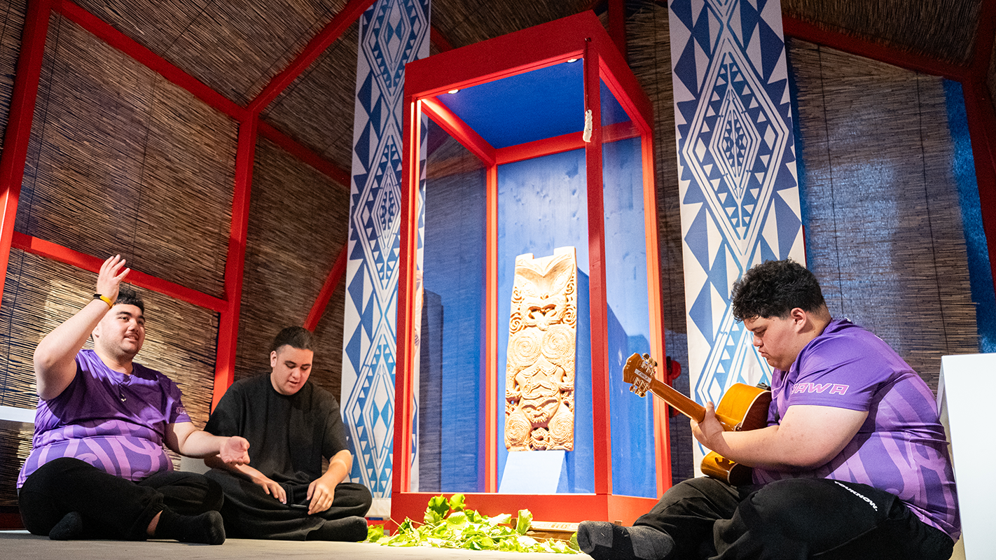 Drei Mitglieder der Māori-Delegation sitzen mit einer Gitarre um die erleuchtete Vitrine mit Hinematioros Pou